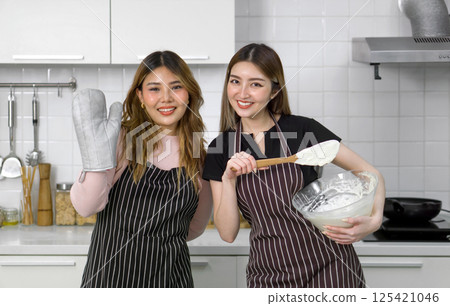 Two women in apron smiling in the kitchen, one holding an oven mitt, the other mixing batter in a bowl. Two women in apron smiling in the kitchen, one holding an oven mitt, the other mixing batter in a bowl. 125421046