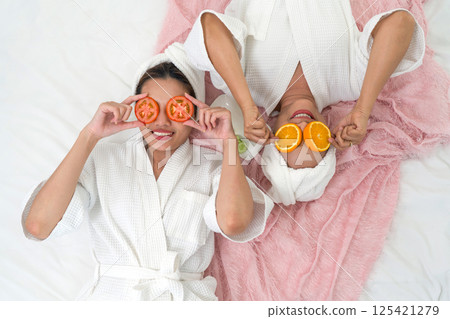 Two women in white robes are lying on a pink blanket, playfully holding slice of tomato and orange over their eye. They are at a spa, enjoying a day of relaxation and pampering. 125421279