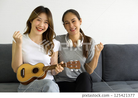 Two asian women playing Ukulele guitar, sitting on sofa in the living room. Enjoy leisure weekend at home. Stress free concept. 125421377