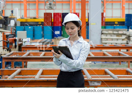 A dedicated female engineer dressed in safety gear holding tablet computer amidst a sprawling factory. A dedicated female engineer dressed in safety gear holding tablet computer amidst a sprawling factory. 125421475