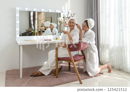 Asian mother and daughter share a joyful moment as they apply skincare product with a brush in cozy home spa. They are dressed in comfortable robe and towel, sit in front of a vanity mirror with light Asian mother and daughter share a joyful moment as they apply skincare product with a brush in cozy home spa. They are dressed in comfortable robe and towel, sit in front of a vanity mirror with light 125421512
