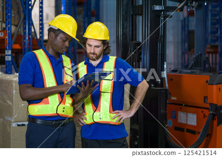 Two warehouse worker in yellow hard hat and safety vest discuss operational detail while examining document in an industrial setting. They focus intently on their task. 125421514
