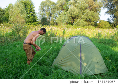 The man installs and assembles tourist tent in a camping in nature in the forest. Domestic tourism, active summer holidays, family adventures. Ecotourism, sport, hike The man installs and assembles tourist tent in a camping in nature in the forest. Domestic tourism, active summer holidays, family adventures. Ecotourism, sport, hike 125425067