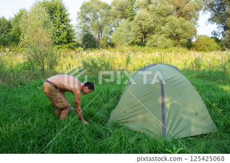 The man installs and assembles tourist tent in a camping in nature in the forest. Domestic tourism, active summer holidays, family adventures. Ecotourism, sport, hike The man installs and assembles tourist tent in a camping in nature in the forest. Domestic tourism, active summer holidays, family adventures. Ecotourism, sport, hike 125425068