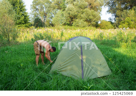 The man installs and assembles tourist tent in a camping in nature in the forest. Domestic tourism, active summer holidays, family adventures. Ecotourism, sport, hike The man installs and assembles tourist tent in a camping in nature in the forest. Domestic tourism, active summer holidays, family adventures. Ecotourism, sport, hike 125425069