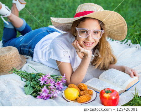 Woman in sunhat reading on picnic blanket, flowers and snacks, cheerful summer relaxation 125426091