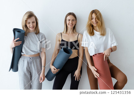 A group of young sports girls with yoga mats standing against a white wall. Girlfriends in the gym relaxing after fitness or yoga, indoors 125431356