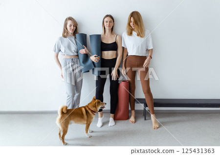 Beautiful young girls in their hands with a yoga mat or karemat stand against the wall smiling before training, and with them a dog indoors Beautiful young girls in their hands with a yoga mat or karemat stand against the wall smiling before training, and with them a dog indoors 125431363