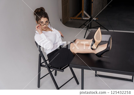Stylish woman in white blouse and black skirt resting legs on table in studio Stylish woman in white blouse and black skirt resting legs on table in studio 125432258