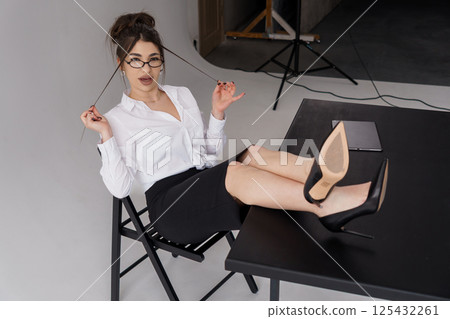 Sophisticated young woman in white shirt and black skirt playfully crosses legs on chair in studio Sophisticated young woman in white shirt and black skirt playfully crosses legs on chair in studio 125432261