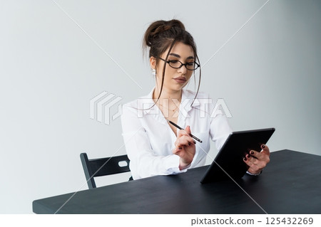 Focused young woman working on tablet with stylus at dark desk, wearing glasses and white shirt 125432269