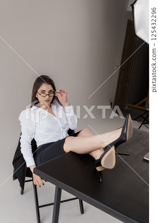 Stylish businesswoman playfully poses with legs on desk, wearing a white shirt and black pencil skirt in a studio setting 125432296