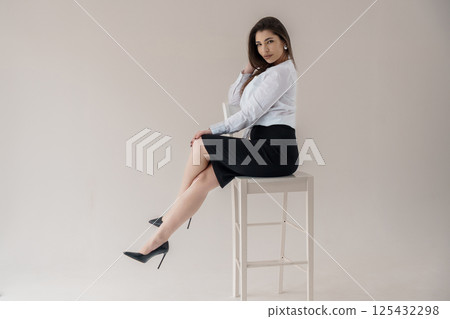 Elegant woman in white shirt and black skirt sitting on stool, posing in studio 125432298