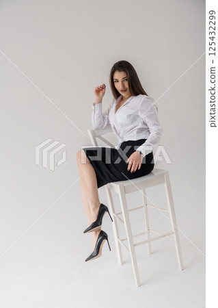 Elegant businesswoman sitting on a white chair in a formal outfit, gazing directly at camera 125432299