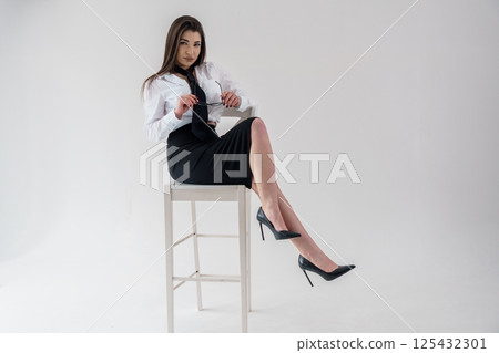 Elegant businesswoman sitting on stool with legs crossed, holding glasses, white background 125432301