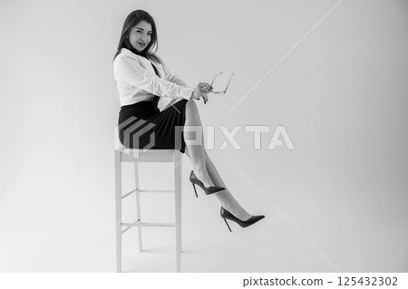 Stylish businesswoman sitting confidently on stool, holding glasses, black and white portrait Stylish businesswoman sitting confidently on stool, holding glasses, black and white portrait 125432302