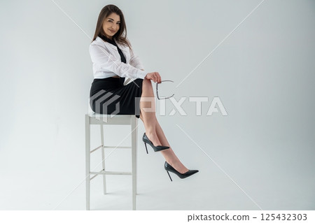 Confident businesswoman sitting on stool holding glasses, professional attire, studio shot 125432303