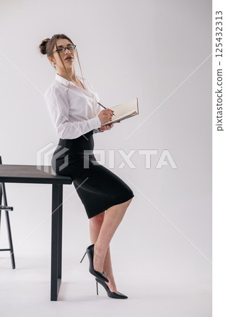 Elegant businesswoman standing near desk writing in notepad, professional attire, confident pose Elegant businesswoman standing near desk writing in notepad, professional attire, confident pose 125432313