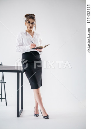Elegant businesswoman writing in a notebook while leaning on a desk in a modern office setting Elegant businesswoman writing in a notebook while leaning on a desk in a modern office setting 125432315