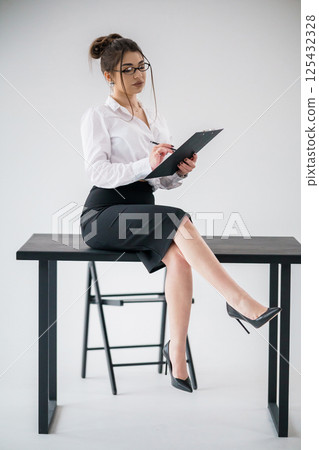 Elegant businesswoman sitting on table writing on clipboard in modern office environment 125432328