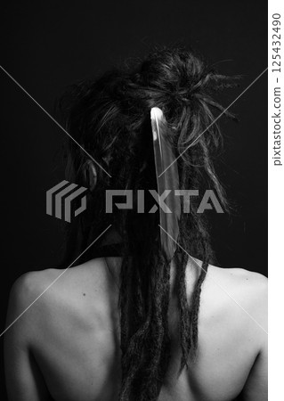 Studio portrait of woman with long dreadlocks and big feather in her hair standing back to camera 125432490