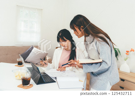 Female higher education student using laptop and studying book in home, Online learning. 125432711