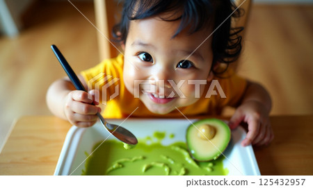 Smiling Pacific Islander baby joyfully creating art with avocado on a tray during a sunny morning 125432957