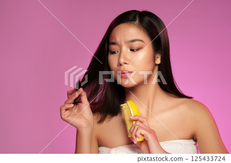 Woman styling her hair with a comb while looking at it against a pink background in a beauty-focused setting 125433724