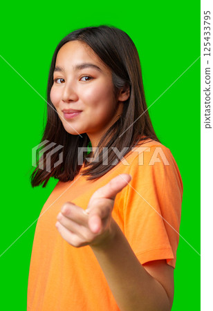 Young woman wearing an orange shirt poses confidently against a bright green background inviting viewers to engage 125433795