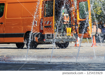 Orange utility service van doors open parked near working fountain in city street center. Fountain system maintenance, inspection winterization work. Equipment tools water accident emergency  vehicle 125433929