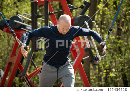 Adult sporty man exercising on resistance machine at outdoor public gym park. Fitness coach show and make exercise. Urban sports ground Adult sporty man exercising on resistance machine at outdoor public gym park. Fitness coach show and make exercise. Urban sports ground 125434528