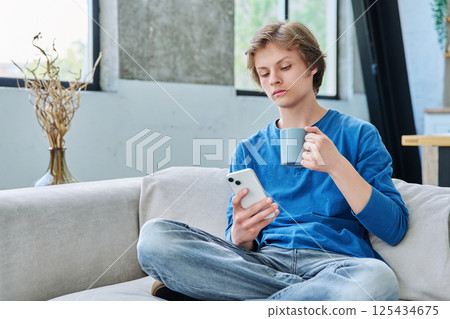 Relaxed teenager guy sitting at home on the couch with smartphone and cup of tea 125434675