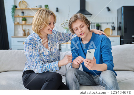 Middle aged mother and teenage son sitting together at home looking at smartphone 125434678