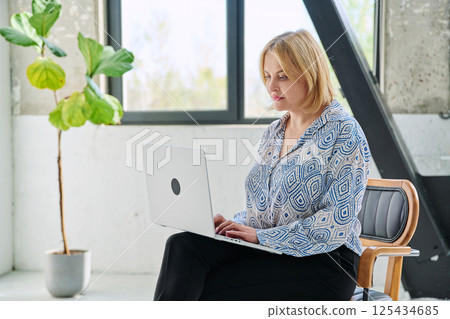 Serious middle aged business woman sitting in chair typing on laptop computer 125434685