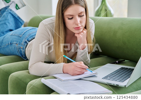 Young student girl lying at home on couch with laptop, notebook, textbook Young student girl lying at home on couch with laptop, notebook, textbook 125434880