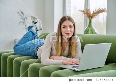 Portrait of young woman lying on sofa at home with laptop looking at camera Portrait of young woman lying on sofa at home with laptop looking at camera 125434883