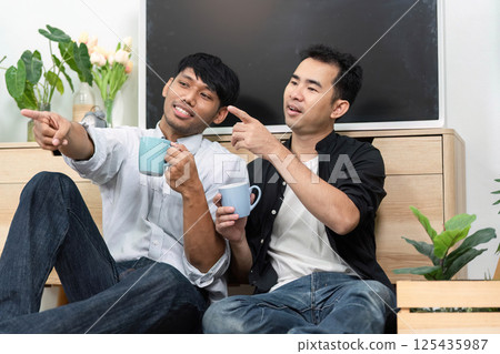 Connection and LGBT Love. Two men sharing a joyful moment over coffee, pointing and laughing together in a cozy living room. Connection and LGBT Love. Two men sharing a joyful moment over coffee, pointing and laughing together in a cozy living room. 125435987