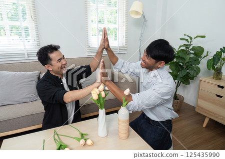 Playfulness and LGBT Joy. Two men engaging in a fun high-five, celebrating their connection in a bright living space. 125435990