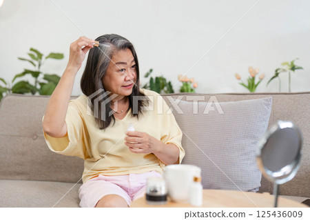 Beauty and Age Diversity. A senior woman applying hair serum with confidence in her living room. 125436009
