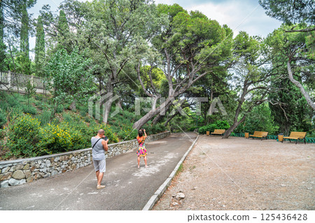 Man and woman taking photos in a scenic park with leaning trees, stone wall, benches, and lush greenery on a cloudy day 125436428