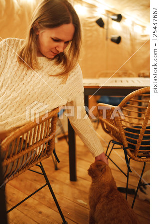 A woman in a white sweater strokes a ginger cat in a cafe in the evening. A woman in a white sweater strokes a ginger cat in a cafe in the evening. 125437662
