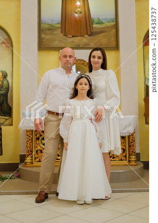 A festive family stands in a bright church. A girl in a long dress beside her parents radiates calm and spirituality. A festive family stands in a bright church. A girl in a long dress beside her parents radiates calm and spirituality. 125438737