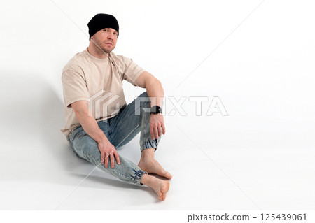 Man in beige t-shirt and black beanie sitting barefoot Man in beige t-shirt and black beanie sitting barefoot 125439061