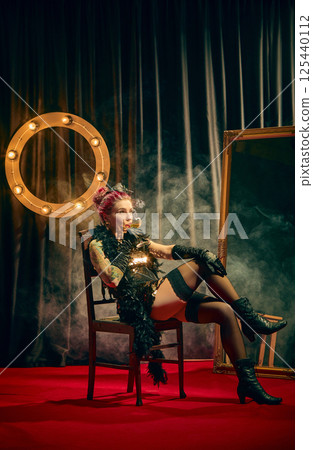 Tattooed cabaret woman sipping drink in seductive mood with feather boa and moody smoke against vintage aesthetic theater background. Tattooed cabaret woman sipping drink in seductive mood with feather boa and moody smoke against vintage aesthetic theater background. 125440112