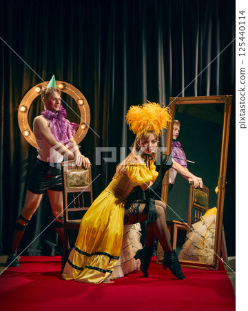 Flirty cabaret duo, elegant woman in yellow dress with cigarette and young guy in short, striking theatrical pose near mirror with vintage chairs. Flirty cabaret duo, elegant woman in yellow dress with cigarette and young guy in short, striking theatrical pose near mirror with vintage chairs. 125440114