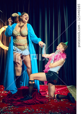 Cabaret queen, overweight woman in blue robe teasing young man with party hat and glitter shorts in stage comedy. 125440165