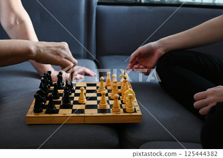 A grandmother and a teenage granddaughter are enthusiastically playing chess on a cozy sofa, demonstrating strategy and concentration in a cozy living room environment 125444382
