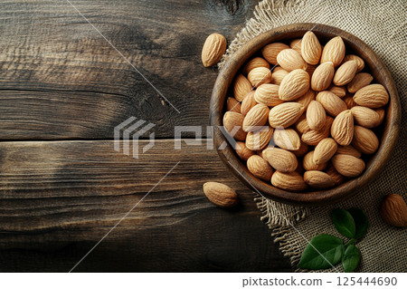 A wooden bowl filled with whole, unpeeled almonds placed on a textured fabric surface, showcasing their natural, rustic appearance in a close-up view. 125444690
