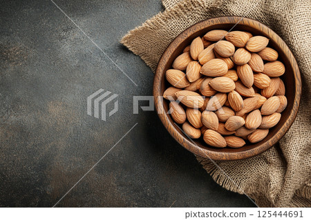 A wooden bowl filled with whole, unpeeled almonds placed on a textured fabric surface, showcasing their natural, rustic appearance in a close-up view. 125444691