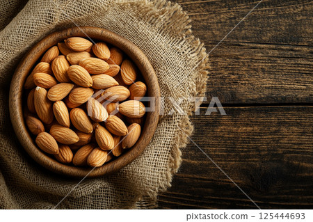 A wooden bowl filled with whole, unpeeled almonds placed on a textured fabric surface, showcasing their natural, rustic appearance in a close-up view. 125444693